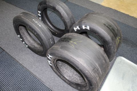 2003 Ford Crown Victoria - Four drag racing slicks marked with white "M H" lettering and faint yellow marks arranged on a gray floor.
