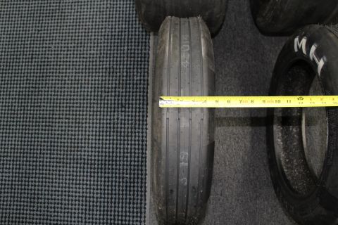 2003 Ford Crown Victoria - narrow ribbed front-runner drag tire being measured across its width with a yellow tape measure on a gray floor.