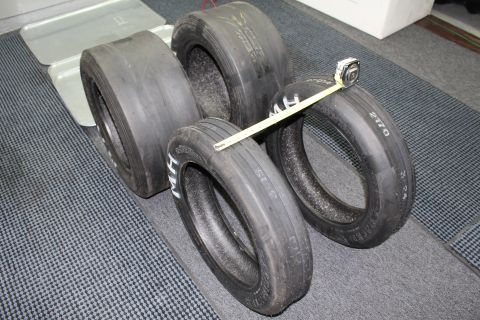 2003 Ford Crown Victoria - Four drag racing tires arranged on a gray floor with a yellow tape measure laid diagonally across them.
