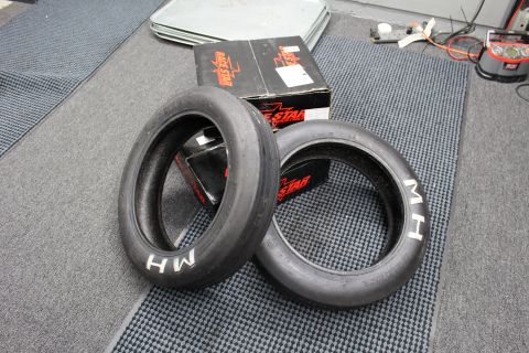 2003 Ford Crown Victoria - Two M/H drag slick tires lie flat on a floor mat beside black Race Star boxes in a workshop.