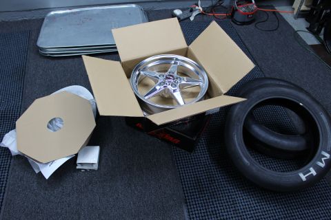 2003 Ford Crown Victoria - polished five-spoke drag wheel sits in an open cardboard box beside octagonal cardboard inserts and an M/H tire.