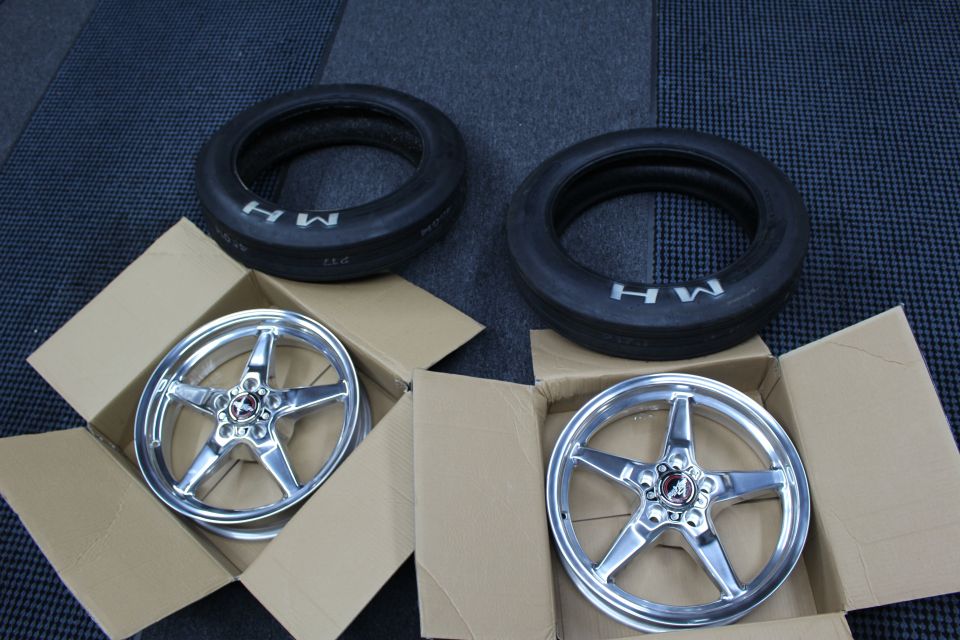 Two polished five-spoke drag wheels in boxes beside two MH front-runner tires