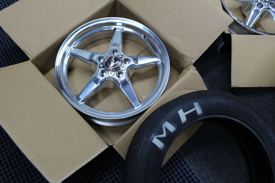Close-up of a polished five-spoke drag wheel in a box beside an MH tire