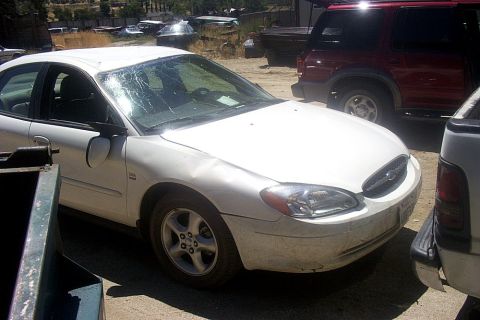 2000 Ford Taurus - A white 2000 Ford Taurus sedan with visible damage sits in a junkyard next to other vehicles.