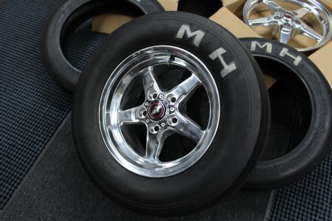 2003 Ford Crown Victoria - polished five-spoke drag wheel mounted on an MH slick sits among loose MH tires and other wheels on a dark mat.