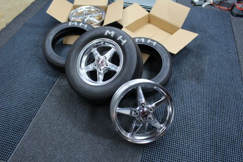 2003 Ford Crown Victoria - Two polished five-spoke wheels mounted on MH drag slicks rest among loose tires and cardboard boxes.