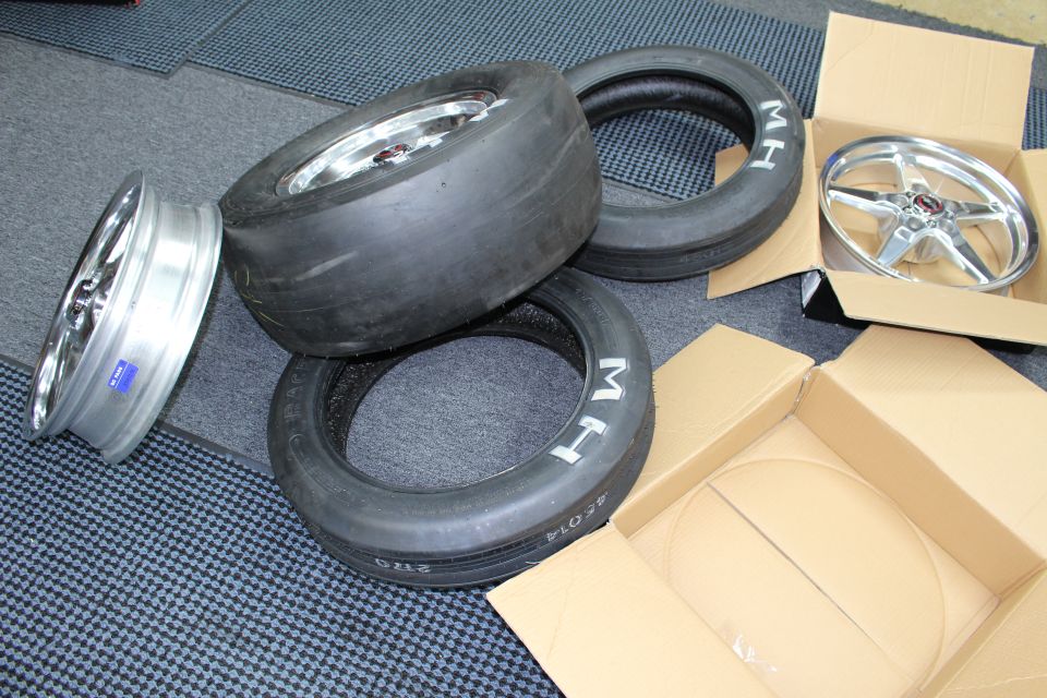 Stacked drag slicks and skinny MH tires with polished wheels on a dark mat