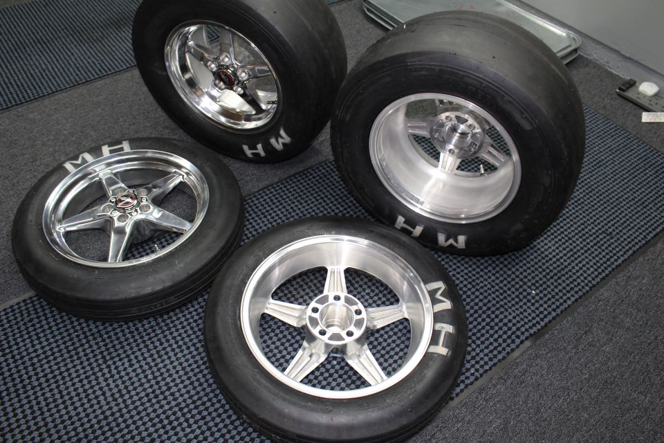 Four polished five-spoke drag wheels with mounted M&H slick tires on a checkered floor