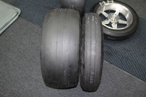 2003 Ford Crown Victoria - Two drag slick tires stood upright side by side showing a noticeable width difference, a mounted wheel behind on a checkered floor.