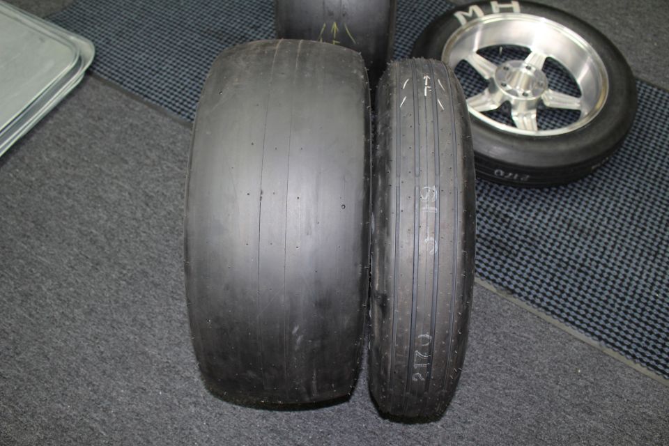 Two slick tires standing side by side showing tread-width difference on a checkered floor