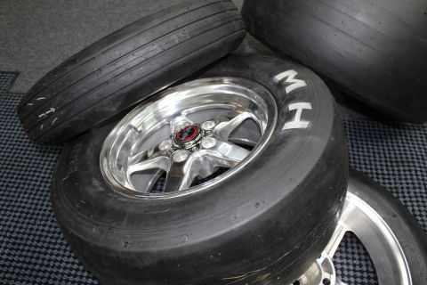 2003 Ford Crown Victoria - polished five-spoke wheel on a drag slick with white MH lettering rests among other tires on a mat.