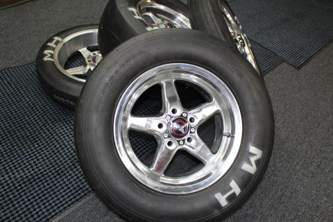 2003 Ford Crown Victoria - polished five-spoke drag wheel with a red center cap and MH-lettered slick stands beside other wheels.