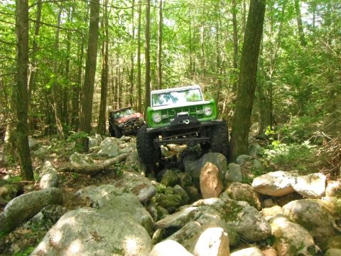 1988 Ford Bronco - Two off-road vehicles tackle a challenging path through a dense, rocky forest.