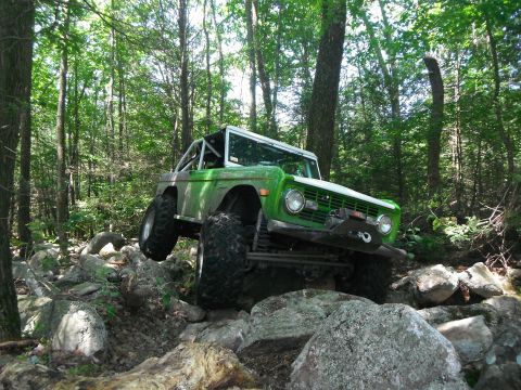 1988 Ford Bronco - Off-road SUV with a roll cage tackles a challenging rocky terrain in a wooded area.