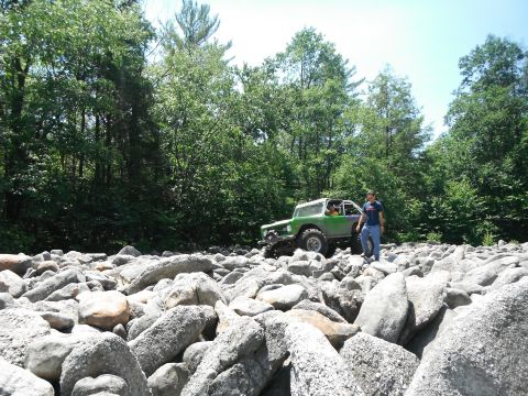 1988 Ford Bronco - A modified green off-road vehicle traverses a rocky terrain with a person standing nearby.