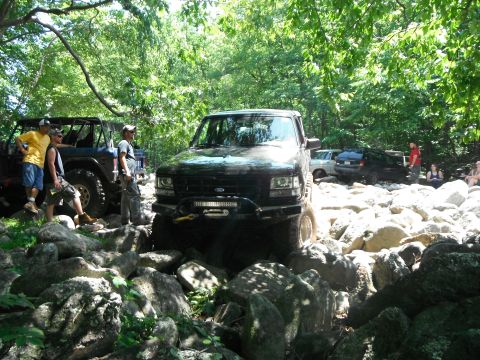 1988 Ford Bronco - Several off-road vehicles traverse a boulder-strewn path under a canopy of trees.
