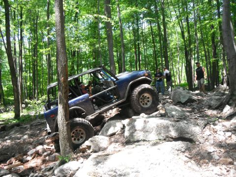1988 Ford Bronco - A blue off-road vehicle navigates a challenging rocky trail through a dense forest with several people watching.