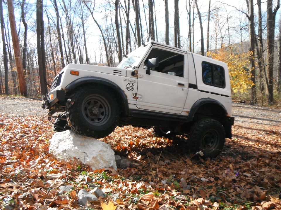 White 1991 Suzuki Samurai climbing over a large rock on a wooded trail