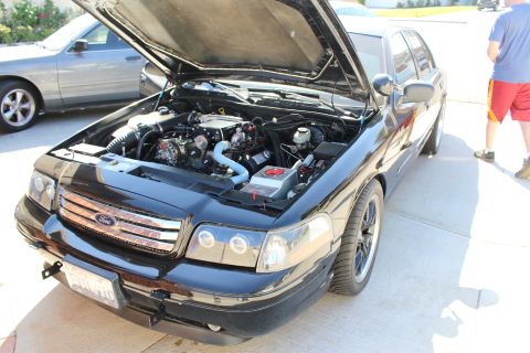 2003 Ford Crown Victoria - black Ford sedan with its hood raised sits in a driveway, exposing the engine bay, with people standing nearby.