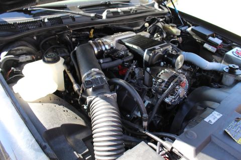 2003 Ford Crown Victoria - Open engine compartment showing a black ribbed intake hose, white coolant reservoir, battery and supercharger assembly under a raised hood.