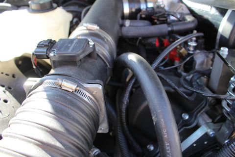 2003 Ford Crown Victoria - Close-up of a black mass-air-flow housing on a rubber intake tube secured by a band clamp, with braided hoses behind in the engine bay.
