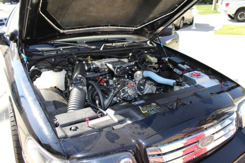 2003 Ford Crown Victoria - Open engine bay of a dark Ford vehicle with hood raised in a driveway, showing a supercharged motor and a Ford emblem on the front.