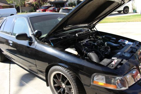 2003 Ford Crown Victoria - black four-door sedan sits in a residential driveway with the hood open, fitted with dark aftermarket wheels.