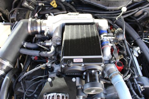 2003 Ford Crown Victoria - black finned Kenne Bell supercharger intercooler with a red oil warning sticker mounted on a dark engine, with blue hoses.