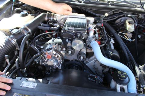 2003 Ford Crown Victoria - engine bay with a Kenne Bell supercharger, chrome alternator and blue coolant pipe; a person's hand rests at the top.