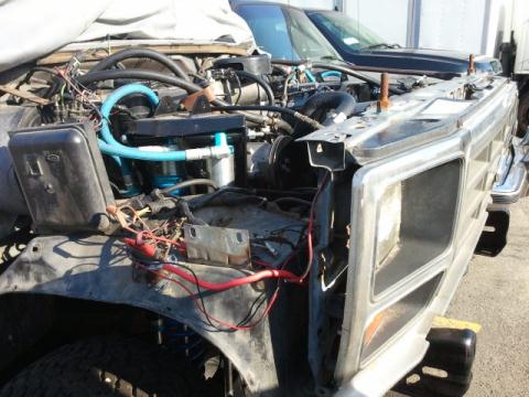 1986 Ford Bronco - The engine bay of a silver Ford Bronco shows exposed wiring and blue hoses, with another vehicle visible behind it.