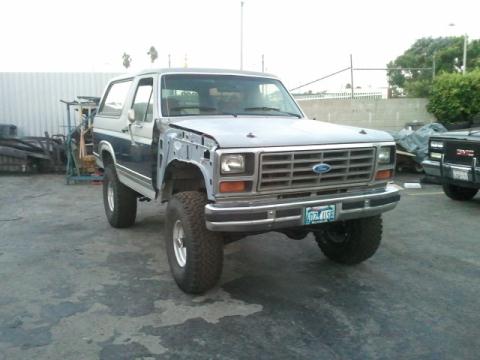 1986 Ford Bronco - A two-tone Ford Bronco sits in a lot with its front fender missing and a second vehicle visible in the background.