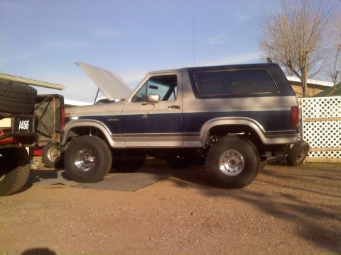 1986 Ford Bronco - Side view of a lifted 1986 Ford Bronco with its hood open, parked on dirt.
