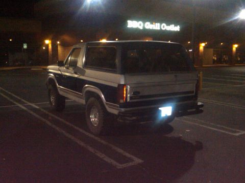 1986 Ford Bronco - A two-tone dark blue and silver Ford Bronco is parked in a lot at night, with a sign for BBQ Grill Outlet visible.
