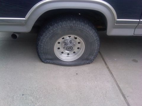 1986 Ford Bronco - A flat tire is visible on the side of a 1986 Ford Bronco.