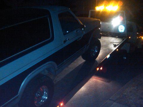 1986 Ford Bronco - A tow truck is loading a Ford Bronco onto its flatbed in the dark.