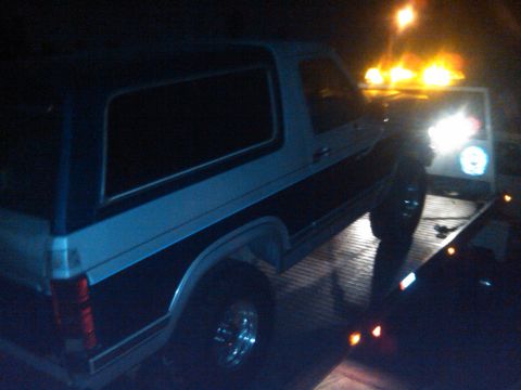 1986 Ford Bronco - A Ford Bronco is loaded onto a tow truck under the glow of emergency lights.