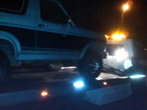 1986 Ford Bronco - A 1986 Ford Bronco is loaded onto a tow truck at night, with its hazard lights illuminated.