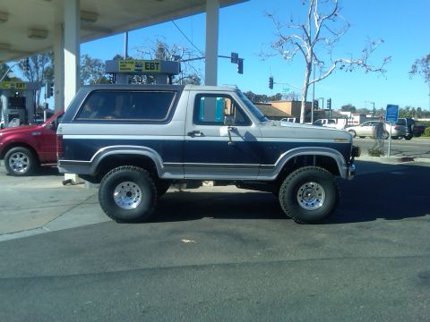 1986 Ford Bronco - A lifted Ford Bronco is parked at a gas station next to a red SUV.