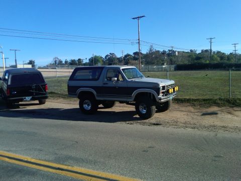 1986 Ford Bronco - Two Ford Broncos are parked side-by-side on a dirt area next to a road under a clear blue sky.