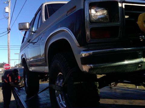 1986 Ford Bronco - Ford Bronco being winched onto a flatbed tow truck with straps around the front tires.