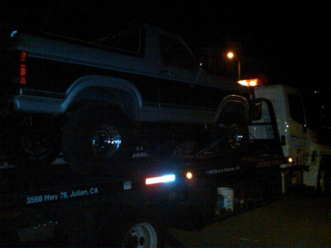 1986 Ford Bronco - A Ford Bronco is transported on a tow truck at night, showing its side profile and taillight detail.