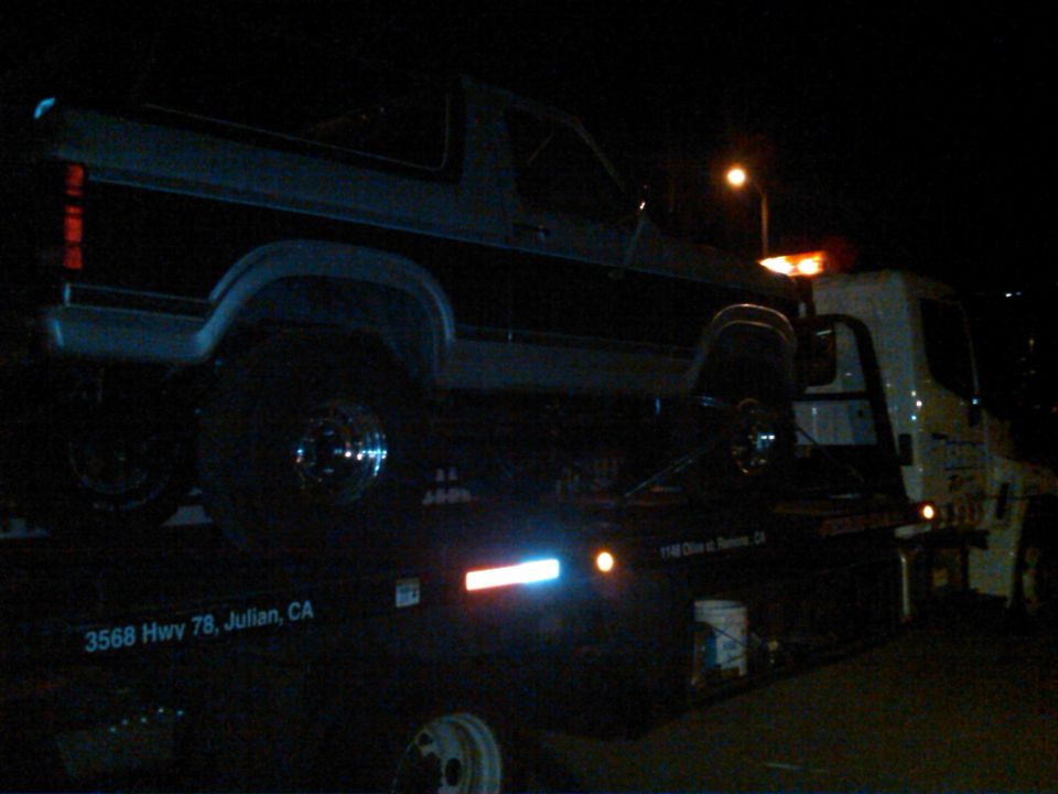 Dark-colored Ford Bronco on a tow truck at night, side view. Visible taillight and fender flares
