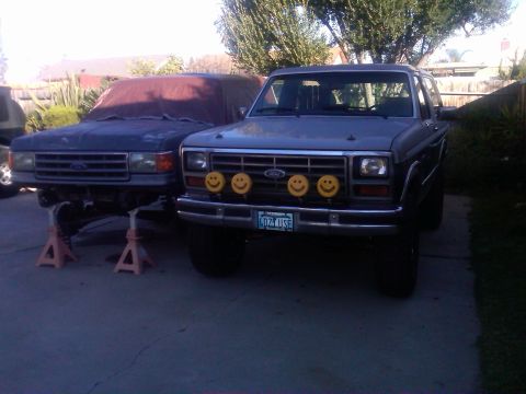 1986 Ford Bronco - Two Ford Broncos are parked in a driveway. The front one has four smiley face auxiliary lights mounted on its bumper.