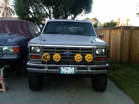 1986 Ford Bronco - Front view of a 1986 Ford Bronco with KC lights and smiley face covers. A second vehicle is parked to the left.