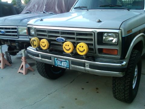 1986 Ford Bronco - Two Ford trucks are parked outside, one with yellow KC lights on its grille.