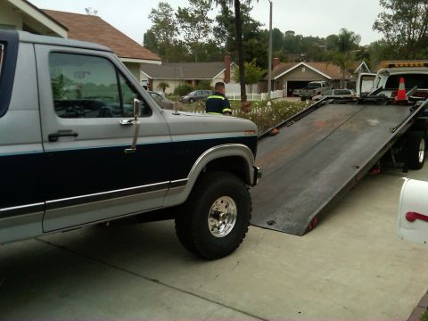 1986 Ford Bronco - A two-tone gray and blue 1986 Ford Bronco is being loaded onto a flatbed tow truck in a residential driveway.
