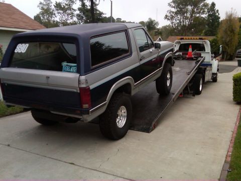 1986 Ford Bronco - Ford Bronco on a flatbed tow truck, viewed from the rear. A tow truck is behind it.