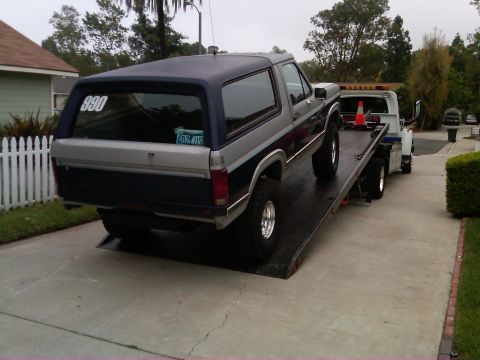 1986 Ford Bronco - A two-tone Ford Bronco is loaded onto a tow truck, with a house and fence in the background.