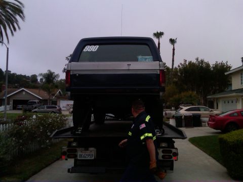 1986 Ford Bronco - 1986 Ford Bronco being loaded onto a tow truck. Tow truck driver visible in foreground.