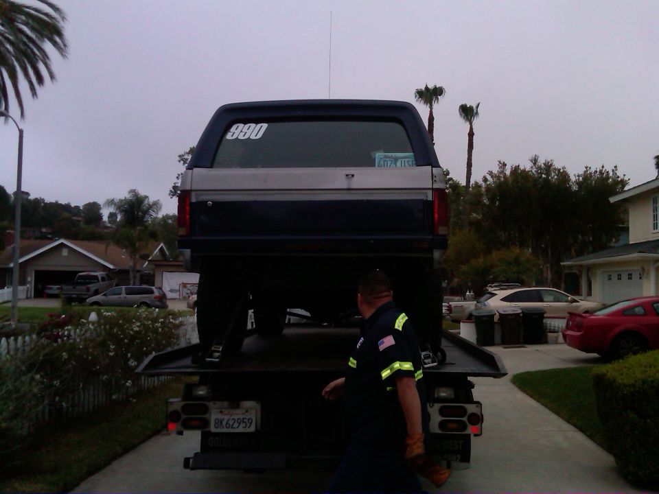 1986 Ford Bronco rear view on a tow truck bed, with a tow truck driver nearby. License plate BK62959